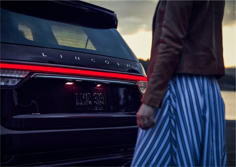 A person is shown near the rear of a 2023 Lincoln Aviator® SUV as the Lincoln Embrace illuminates the rear lights | Parks Lincoln of Tampa in Tampa FL